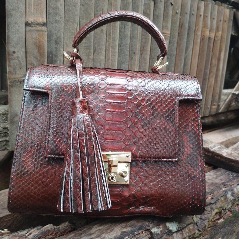 Genuine luxury python leather top-handle handbag featuring polished gold-tone hardware and a matching leather tassel, displayed against a rustic bamboo and wood background