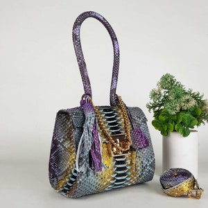 Könnte beinhalten: Eine stilvolle Handtasche mit Schlangenmuster in Grau-, Gold- und Lilatönen. Die Tasche hat einen goldenen Kettenriemen, einen passenden Verschluss und eine dekorative Quaste. Ein passender Gürtel ist ebenfalls sichtbar.