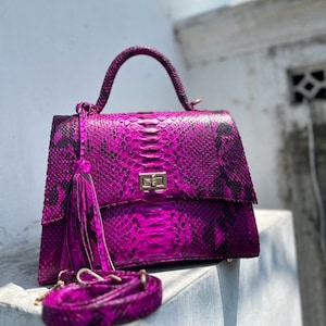 Könnte beinhalten: Eine leuchtend magenta Handtasche mit Schlangenmuster. Die Tasche hat einen oberen Griff, einen abnehmbaren Schulterriemen und eine dekorative Quaste. Der Verschluss ist goldfarben. Die Tasche steht auf einer weißen Oberfläche.