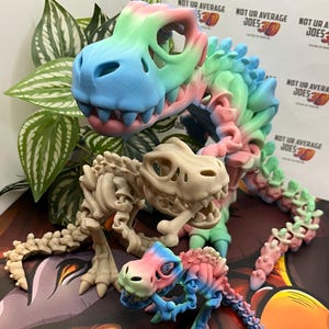May include: Three 3D-printed dinosaur skeletons. One large skeleton is beige, while the other two are smaller and multi-colored with blue, green, pink, and yellow hues. The text "NOT UR AVERAGE JOES 3D" is visible in the background.