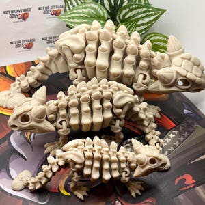 Może przedstawiać: Trzy ruchome szkielety dinozaurów wykonane z białego plastiku. Szkielety są ustawione w zabawny sposób, z największym szkieletem z tyłu i najmniejszym szkieletem z przodu. Szkielety są wszystkie w tej samej pozie, z głową pochyloną na bok i otwartymi ustami.