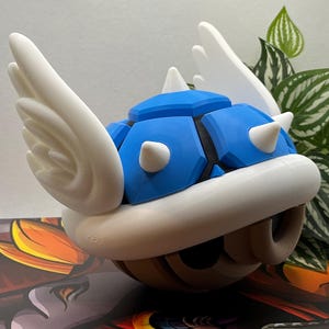 May include: A blue and white 3D printed Super Mario Bros. Koopa shell with wings. The shell is a light blue color with white spikes and white wings. The shell is sitting on a brown surface.