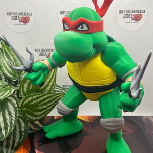 May include: A green 3D-printed Teenage Mutant Ninja Turtle figure, with a red mask and two silver sai. The figure has a yellow plastron and grey accents. The figure is in a fighting stance. The text "NOT UR AVERAGE JOES 3D" is visible.