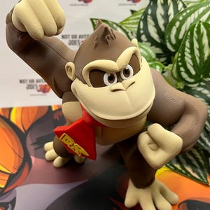 Könnte beinhalten: 3D-gedruckte Donkey Kong-Figur mit braunem Körper, beigem Gesicht und Händen sowie einer roten Krawatte mit der Aufschrift "DK". Die Figur befindet sich in einer dynamischen Pose vor einem farbenfrohen Hintergrund.