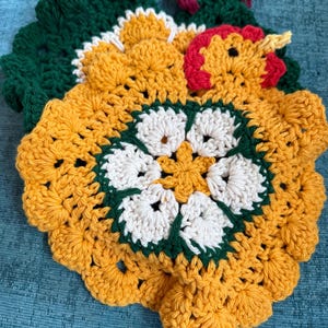 May include: A crochet turkey-shaped potholder featuring a central white flower motif surrounded by golden-yellow yarn petals. The turkey has a dark green tail, a golden-yellow body, a red wattle, and a small yellow beak, all crafted in yarn.