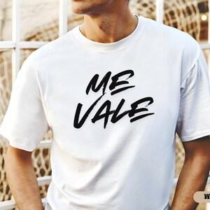 May include: A white t-shirt with the words "ME VALE" in bold, black brush lettering. The shirt is a classic crew neck style and appears to be made of a soft material. The word "WHITE" is in a small, rectangular box in the bottom right corner.