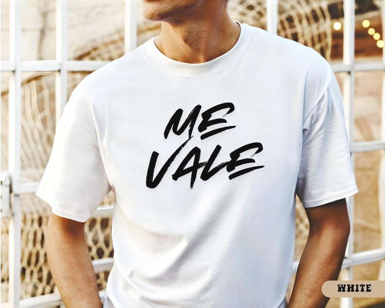 May include: A white t-shirt with the words "ME VALE" in bold, black brush lettering. The shirt is a classic crew neck style and appears to be made of a soft material. The word "WHITE" is in a small, rectangular box in the bottom right corner.