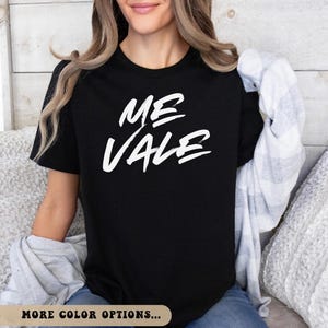 May include: Black t-shirt with the white text "ME VALE" in a handwritten style. The shirt is being worn by a person. The phrase is centered on the front of the shirt. The person is wearing blue jeans and has a gray and white plaid shirt draped over their shoulders.