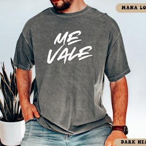 May include: Dark heather gray t-shirt with the white text "ME VALE" in a handwritten style. The shirt is short-sleeved and has a relaxed fit. The text "MANA LOVERS" is at the top right corner and "DARK HEATHER" at the bottom right corner.