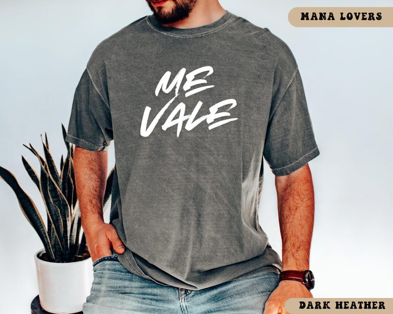 May include: Dark heather gray t-shirt with the white text "ME VALE" in a handwritten style. The shirt is short-sleeved and has a relaxed fit. The text "MANA LOVERS" is at the top right corner and "DARK HEATHER" at the bottom right corner.
