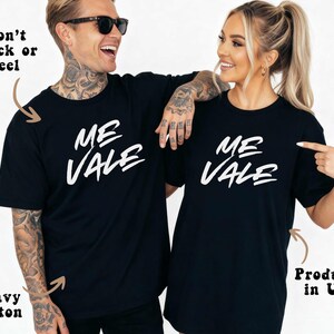 May include: Two people wearing matching black t-shirts with the white text "ME VALE". The shirts are made of heavy cotton and are produced in the USA. The text "Won't Crack or Peel" is also visible.