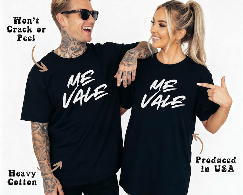 May include: Two people wearing matching black t-shirts with the white text "ME VALE". The shirts are made of heavy cotton and are produced in the USA. The text "Won't Crack or Peel" is also visible.