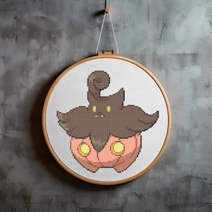Puede incluir: Un patrón de punto de cruz de un personaje Pokémon marrón y rosa con ojos amarillos. El personaje es una criatura parecida a una calabaza con una cabeza marrón y frondosa.