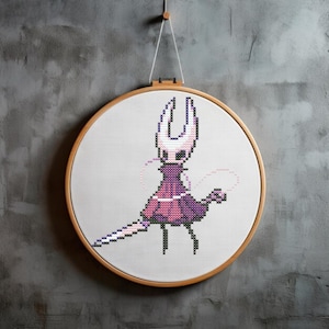 May include: A cross-stitch embroidery hoop with a pixelated image of a pink and purple character with a white mask and long, white ears. The character is holding a white sword and has a pink and purple tail.