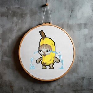 May include: A cross-stitch embroidery hoop with a pixelated image of a crying banana wearing a yellow banana costume. The banana is surrounded by blue pixelated water droplets.