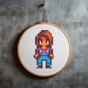 May include: Cross-stitch embroidery hoop with a pixelated image of a person wearing blue overalls and a pink shirt. The person has brown hair and is standing with their arms at their sides.