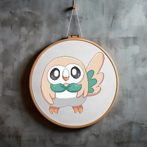 May include: A cross-stitch embroidery hoop with a cartoon image of a brown and white owl with green wings and a green bow tie. The owl has large black eyes and a small orange beak.