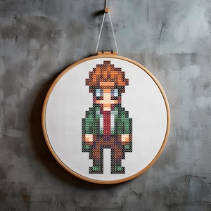 May include: A cross-stitch embroidery hoop with a pixelated image of a person with brown hair, a green jacket, a red tie, brown pants, and green shoes.