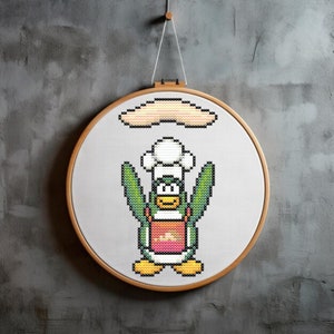 Può includere: Un cerchio da ricamo a punto croce con un pinguino verde che indossa un cappello da chef bianco e tiene una spatola. Il pinguino è in piedi su un tappeto a strisce gialle e arancioni. Una luna crescente gialla è sopra il pinguino.