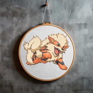 May include: A cross-stitch embroidery of an Arcanine character in a wooden hoop. The artwork features the Pokémon in shades of cream, orange, and black, with an open mouth and a dynamic pose. The hoop is suspended by a white string against a grey wall.