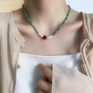 Könnte beinhalten: Eine zarte Halskette mit einer Kette aus kleinen, grünen Perlen. Die Halskette hat ein zentrales Design mit einer roten Perle, umgeben von weißen Perlen. Goldfarbene Akzente sind eingearbeitet, was einen raffinierten und eleganten Look erzeugt.