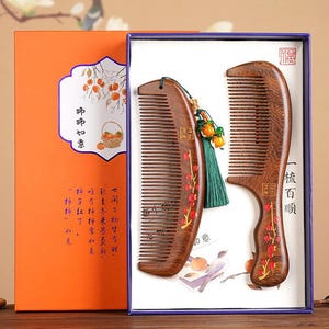 May include: Two wooden combs in a gift box. The combs are decorated with a persimmon design and Chinese characters. The box is orange with a persimmon design and the text "柿柿如意" and "一梳百顺".