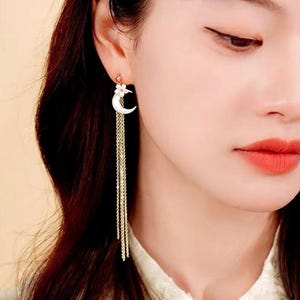 May include: A pair of gold dangle earrings with a crescent moon and flower charm. The earrings have a long chain with multiple strands.