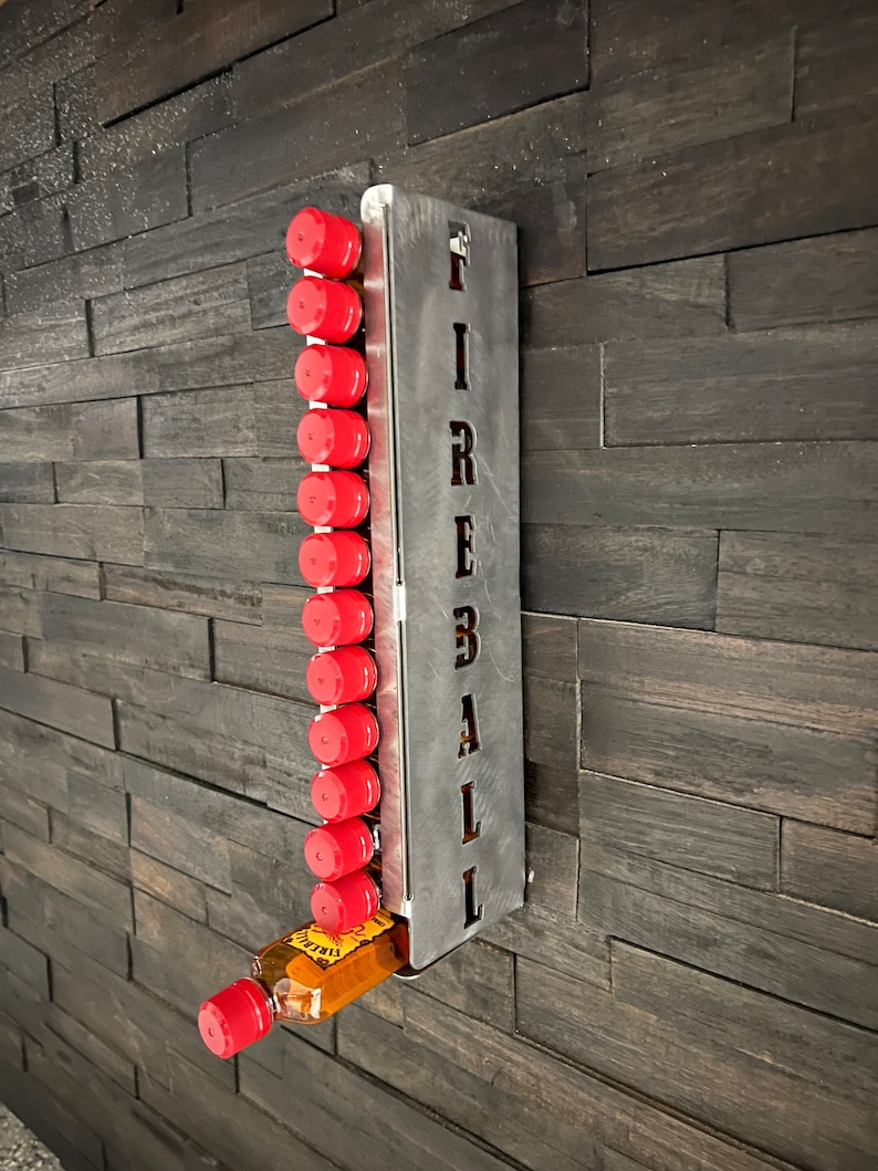Op de afbeelding: Een metalen wandbevestiging met het woord "FIREBALL" uitgesneden, met een rij miniatuurflesjes Fireball whisky. De flesjes zijn rood met zwarte etiketten en de wandbevestiging is zilverkleurig.