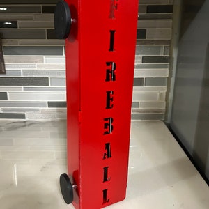 Op de afbeelding: Rode metalen brandkraan met het woord "FIREBALL" uitgesneden in zwarte letters. De brandkraan heeft zwarte wielen aan de onderkant.