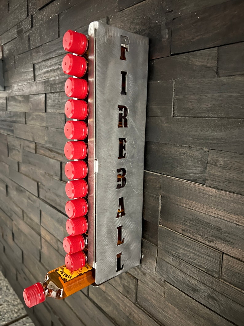 Op de afbeelding: Metalen wandhouder voor flessen met het woord "FIREBALL" uitgesneden. De houder heeft 12 vakken voor flessen en een fles Fireball whisky bevindt zich in het onderste vak.