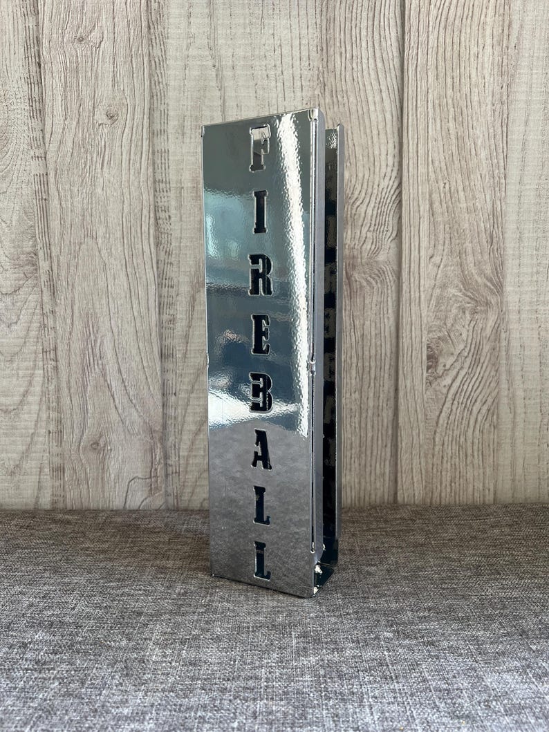 Op de afbeelding: Een zilveren metalen bord met het woord "FIREBALL" uitgesneden in zwarte letters. Het bord staat rechtop op een grijs, getextureerd oppervlak.