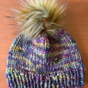 May include: A chunky knit beanie featuring a variegated yarn pattern in shades of purple, blue, yellow, and green. The hat is topped with a large, fluffy faux fur pom-pom in a mix of brown, tan, and cream tones, creating a textured appearance.