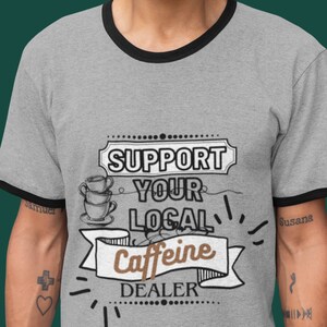 Puede incluir: Una camiseta gris con cuello de canalé negro. La camiseta tiene un diseño gráfico que dice "Apoya a tu distribuidor local de cafeína" con un diseño de taza de café estilizado.