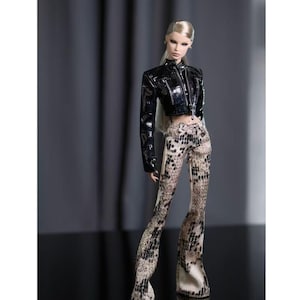 Pode incluir: Uma boneca de moda com cabelo loiro preso num rabo de cavalo baixo, vestindo um casaco preto curto com acabamento brilhante e texturizado de mangas compridas. A boneca também usa calças de cintura subida e corte largo com um padrão de pele de cobra em bege e preto.