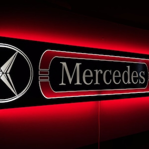 May include: A black and silver sign with the word "Mercedes" in silver lettering. The sign is illuminated with red LED lights and features the Mercedes-Benz logo on either side.