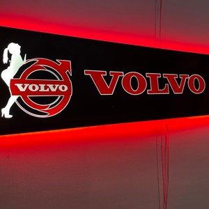 May include: A black sign with red neon trim and the word "VOLVO" in red letters. The sign also features a white silhouette of a woman with the word "VOLVO" in white letters inside a red circle.
