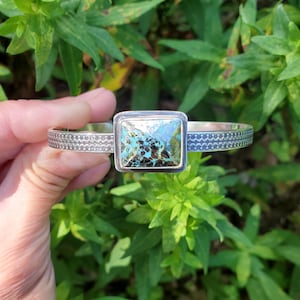 May include: A silver cuff bracelet with a square turquoise stone. The stone is a mix of blue, green, and brown. The bracelet band has a repeating wave pattern and is held by a hand.