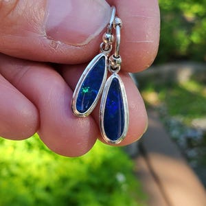 May include: A pair of drop earrings featuring elongated, teardrop-shaped dark blue opals with vibrant green and blue flashes. Each opal is set in a polished silver bezel frame and attached to a small silver hoop component with a spherical bead detail.