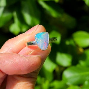 May include: A silver ring featuring a shield-shaped opal gemstone. The opal displays iridescent flashes of blue, green, and pink light. The metal band is polished and smooth, holding the stone in a simple bezel setting that highlights the play of colour.