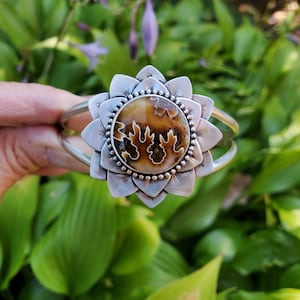 May include: A silver cuff bracelet featuring a large, round, fossilized stone center. The stone is surrounded by a silver flower design with detailed petals. The bracelet is displayed against a backdrop of green foliage.