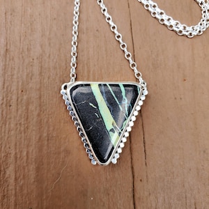 Op de afbeelding: Een zilveren ketting met een driehoekige hanger. De hanger heeft een zwarte steen met groene en gele adering, gezet in een zilveren frame met een kralenrand. De ketting is een fijne zilveren schakel.