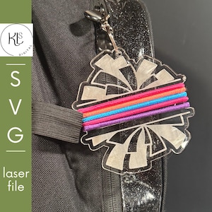 May include: A clear acrylic cheerleading pom-pom keychain featuring a horizontal band of three elastic cords in bright orange, blue, and purple. The keychain is attached to a silver metal lobster clasp and hangs from a black fabric backpack.