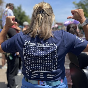 May include: A navy blue t-shirt featuring a Disney Word Search design. The graphic includes a word search puzzle with Disney-themed words and a list of words to find. The shirt is worn by a person with their arms raised.