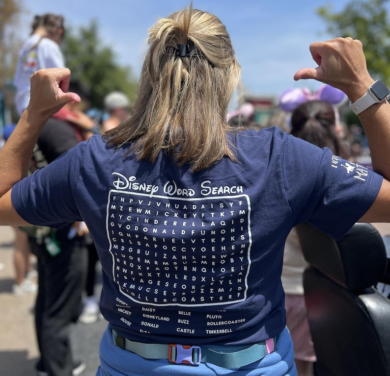 May include: A navy blue t-shirt featuring a Disney Word Search design. The graphic includes a word search puzzle with Disney-themed words and a list of words to find. The shirt is worn by a person with their arms raised.