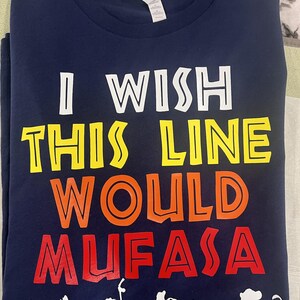May include: A navy blue t-shirt with the phrase "I WISH THIS LINE WOULD MUFASA" in white, yellow, orange, and red. The design includes white cartoon silhouettes of characters from The Lion King at the bottom.