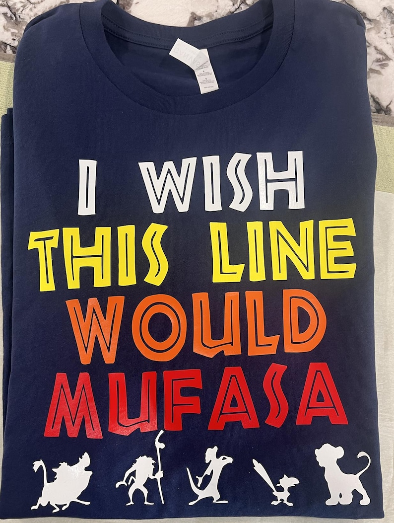 May include: A navy blue t-shirt with the phrase "I WISH THIS LINE WOULD MUFASA" in white, yellow, orange, and red. The design includes white cartoon silhouettes of characters from The Lion King at the bottom.