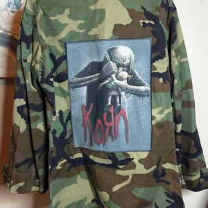 May include: Camouflage military jacket with a large patch featuring a gray figure holding a doll, with the band name "Korn" in red lettering. The jacket has a button-up front, long sleeves, and a classic army green, brown, and black camouflage pattern.