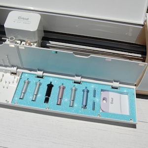 Könnte beinhalten: Eine weiße Cricut-Maschine mit einem Fach, das verschiedene Schneidwerkzeuge in Silber, Schwarz und Blau enthält. Das Fach hat einen hellblauen Glitzerinnenraum, und ein weißer Schaber ist ebenfalls enthalten.