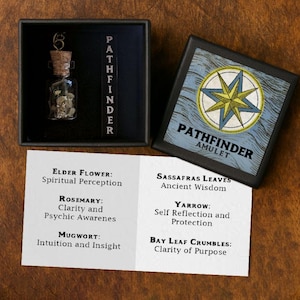 Op de afbeelding: Een Pathfinder-amuletset. De set bevat een kleine glazen fles met een kurk, een zwarte doos met het woord "PATHFINDER" en een kaart met de tekst "ELDER FLOWER: Spiritual Perception". De doos heeft een kompasroosontwerp.