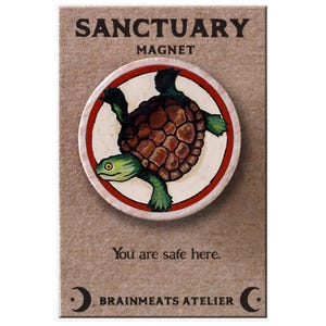 Puede incluir: Un imán redondo con una ilustración de tortuga dentro de un círculo rojo y blanco. El imán lleva la etiqueta "SANCTUARY MAGNET" e incluye el texto "You are safe here." y "BRAINMEATS ATELIER".